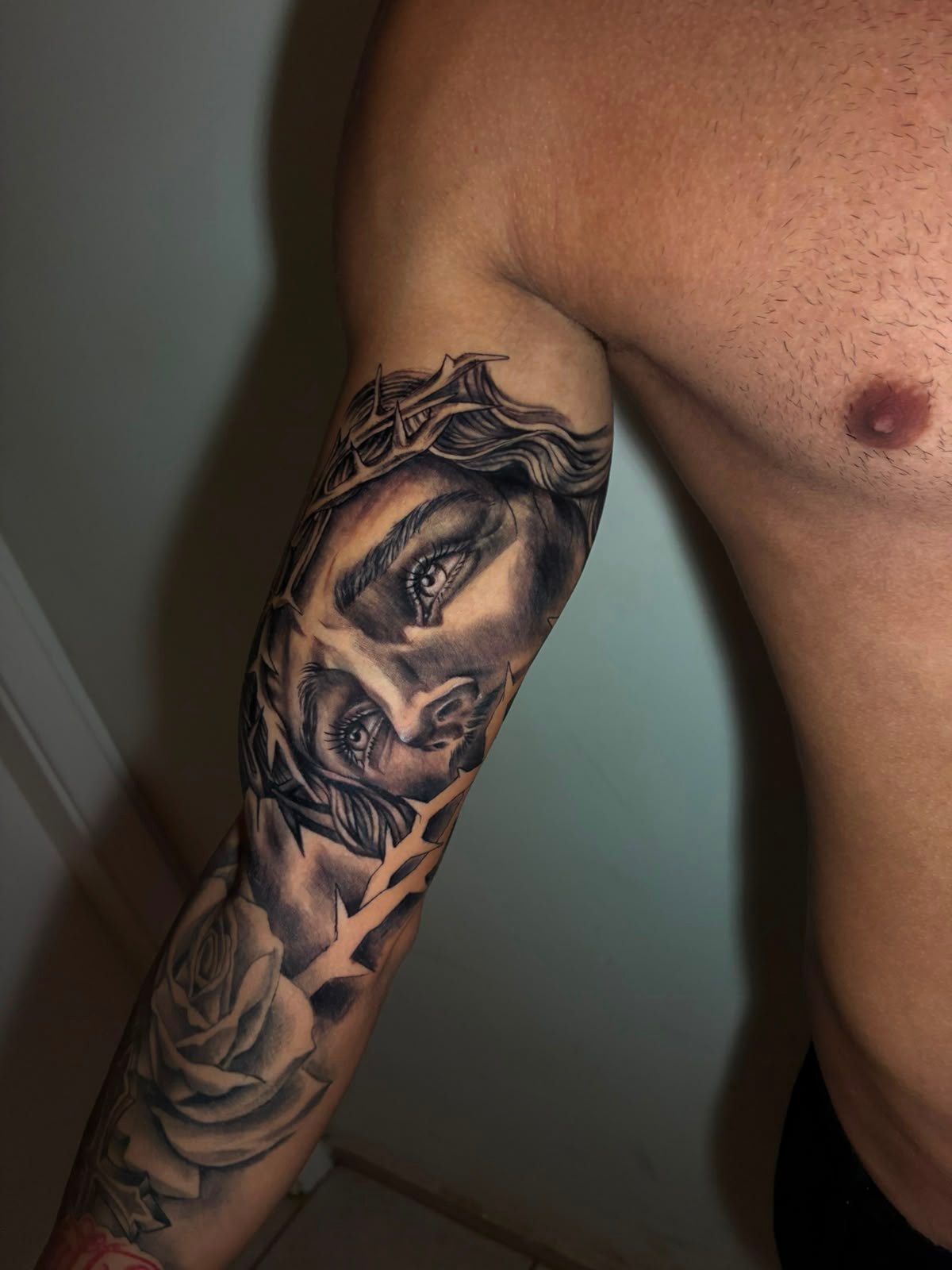 Tatuagem realista de rosto com coroa de espinhos e rosa no braço
